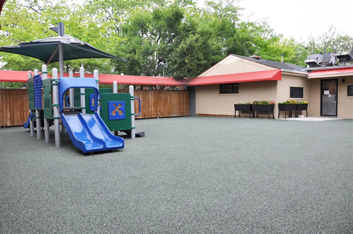 The school's fenced outdoor playground, with a climbing structure, slide, and shaded soft-surface play area